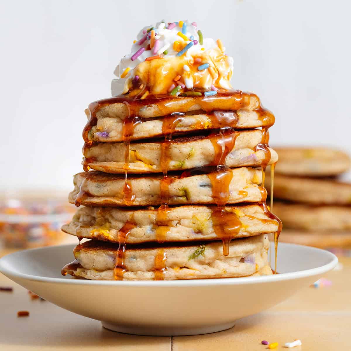 A stack of pancakes with sprinkles with whipped cream and more sprinkles on top and maple syrup dripping down the sides.