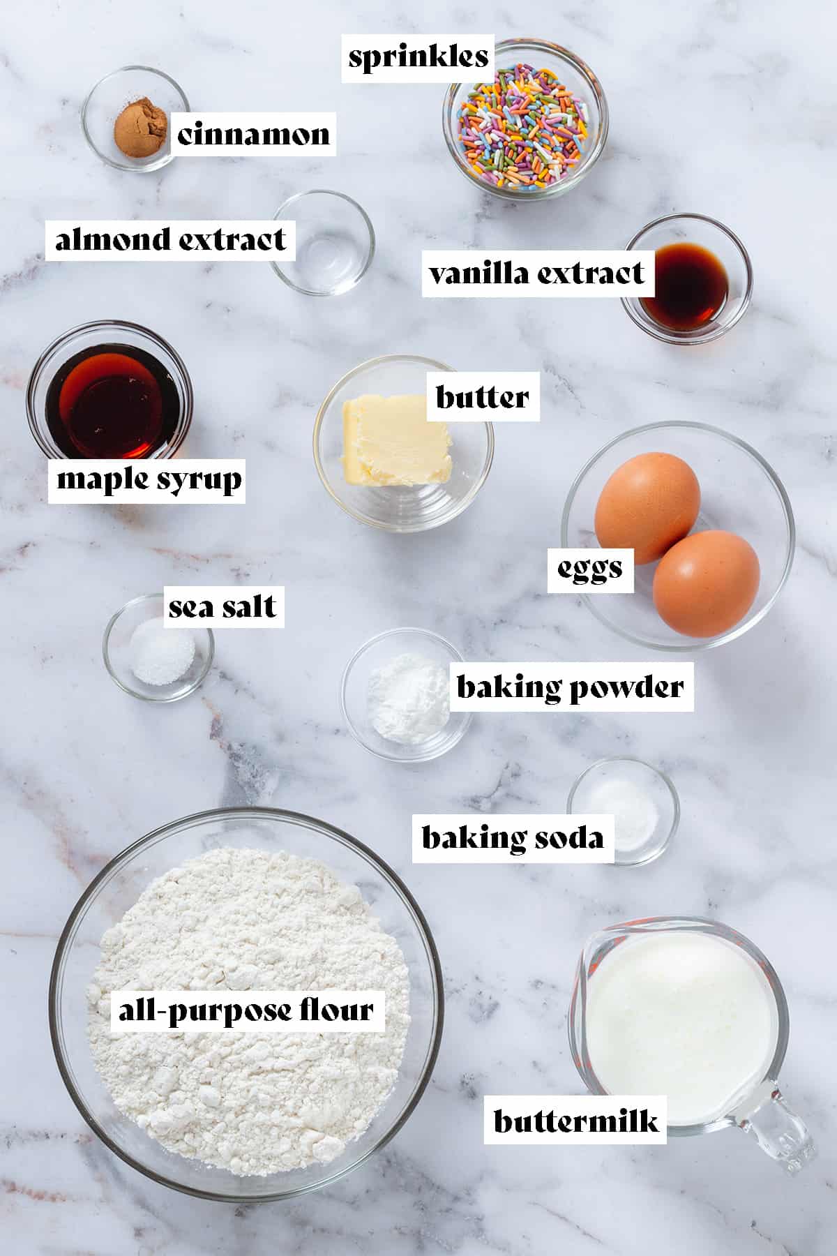 Ingredients like flour, maple syrup, eggs, buttermilk, and butter in glass bowls on a marble background.