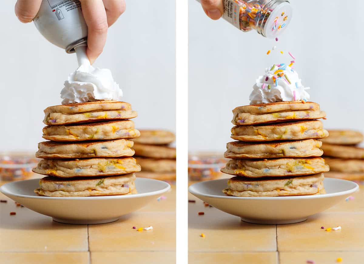 A stack of pancakes with sprinkles being topped with whipped cream and more sprinkles.