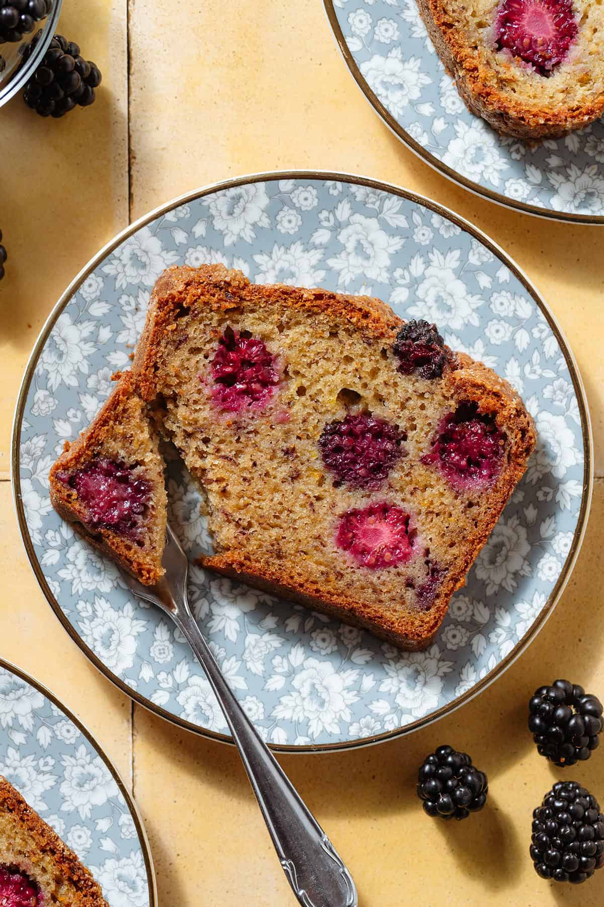 A slice of blackberry banana bread showing multiple blackberries inside on a blue floral plate with a fork cutting off a corner of the slice.