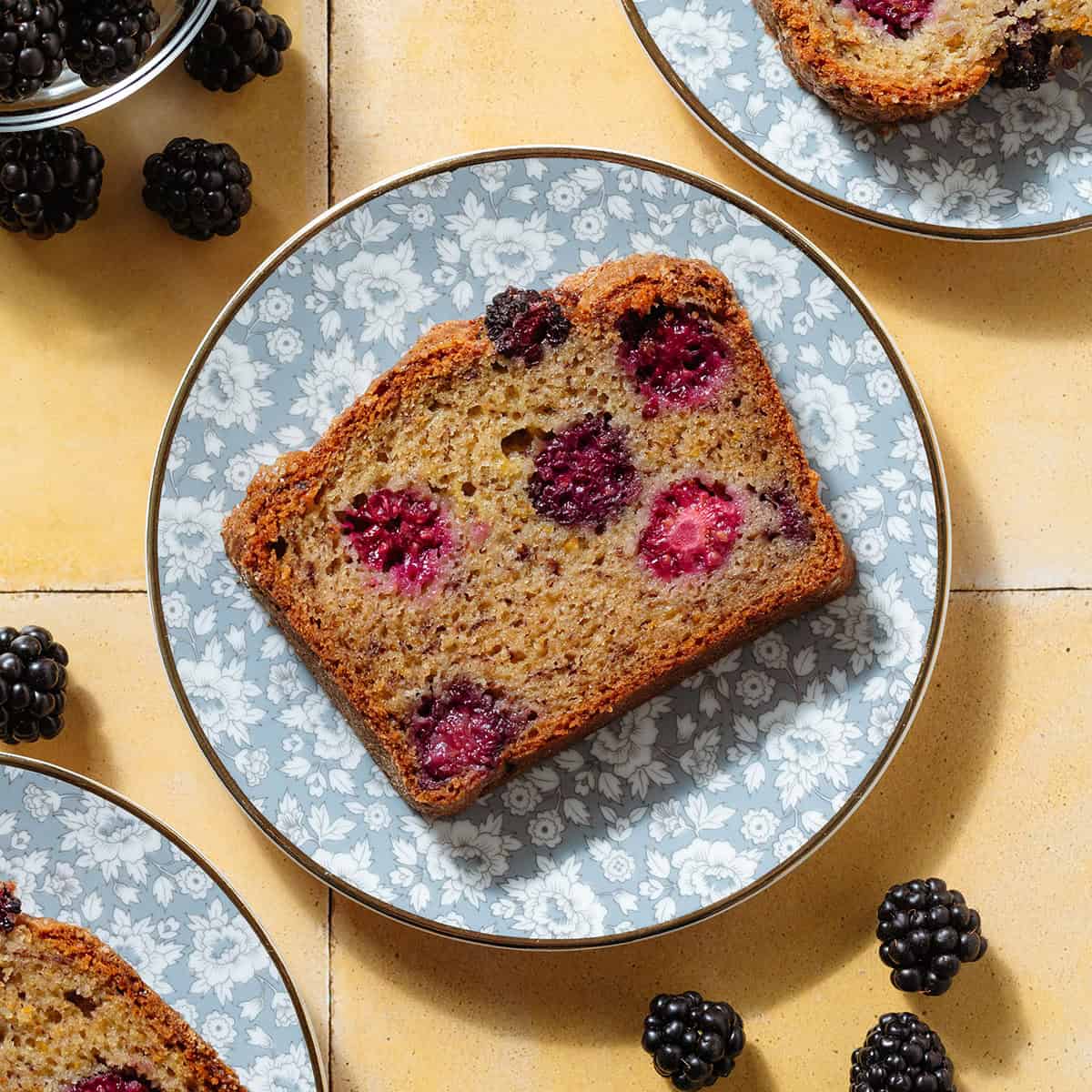 A slice of blackberry banana bread showing multiple blackberries inside on a blue floral plate.