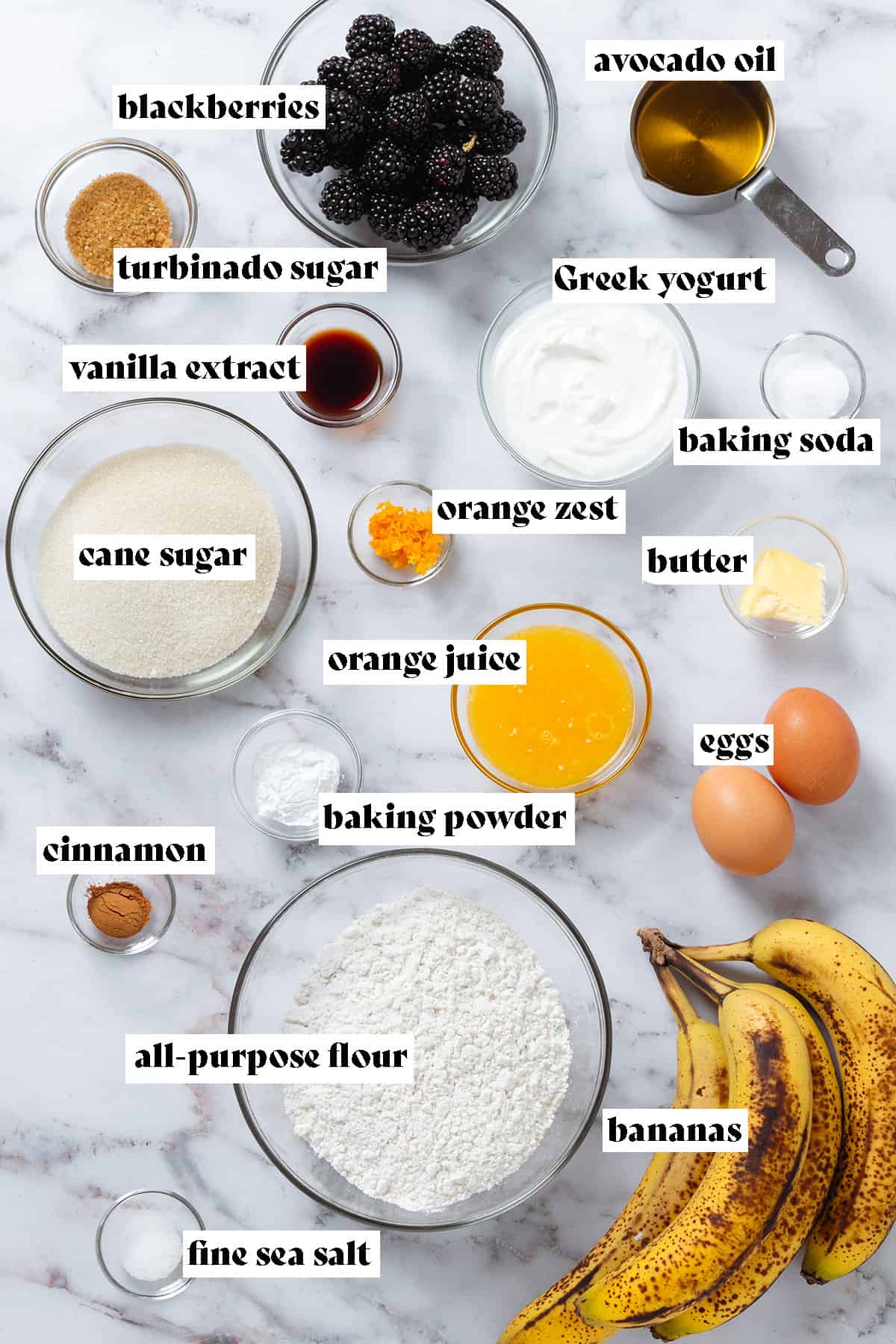 Ingredients in glass bowls like blackberries, flour sugar, yogurt, orange juice and other ingredients all measured and laid out on a marble background with text overlay.