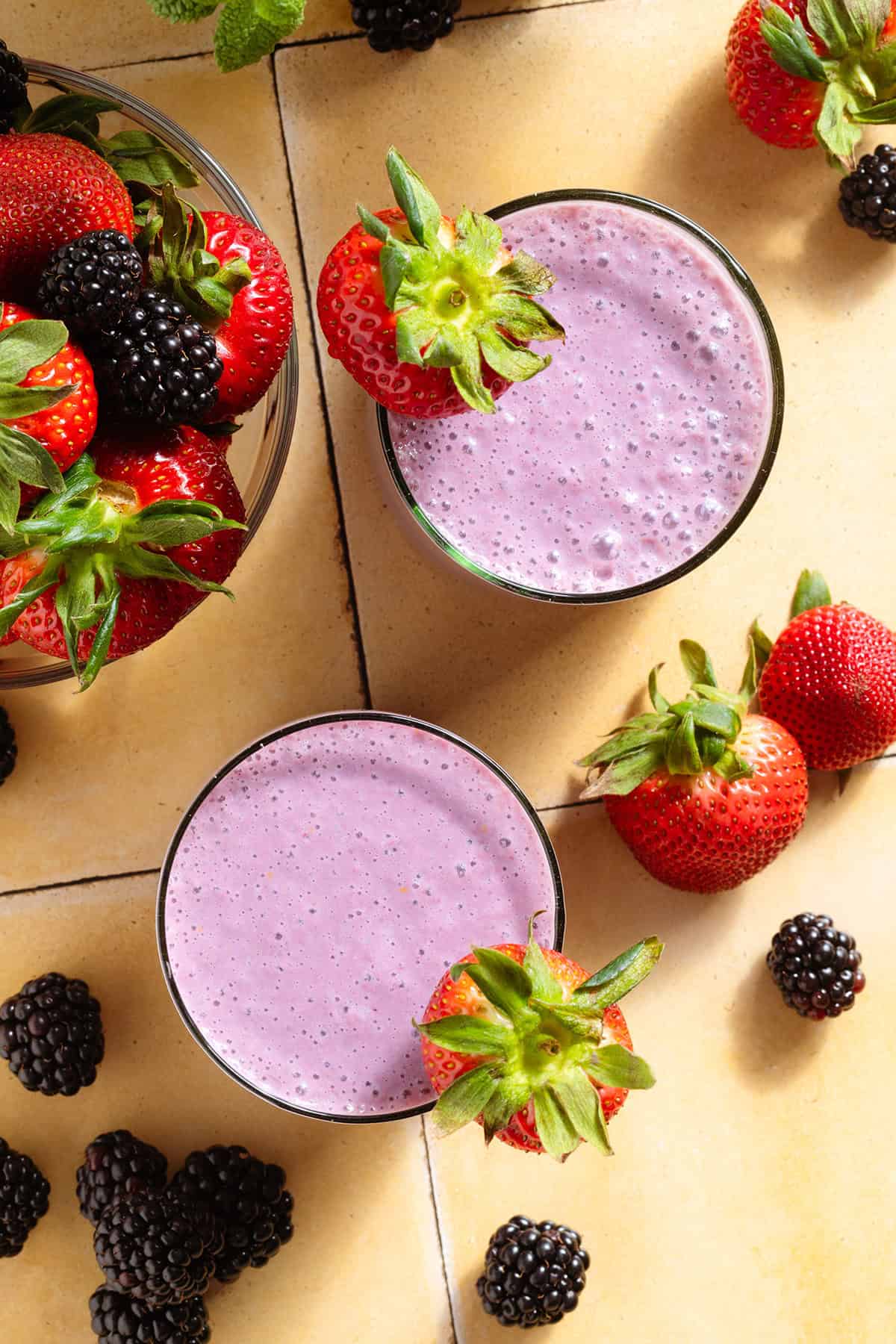 A light purple smoothie in two short glasses garnished with a strawberries and blackberries scattered around the glass.