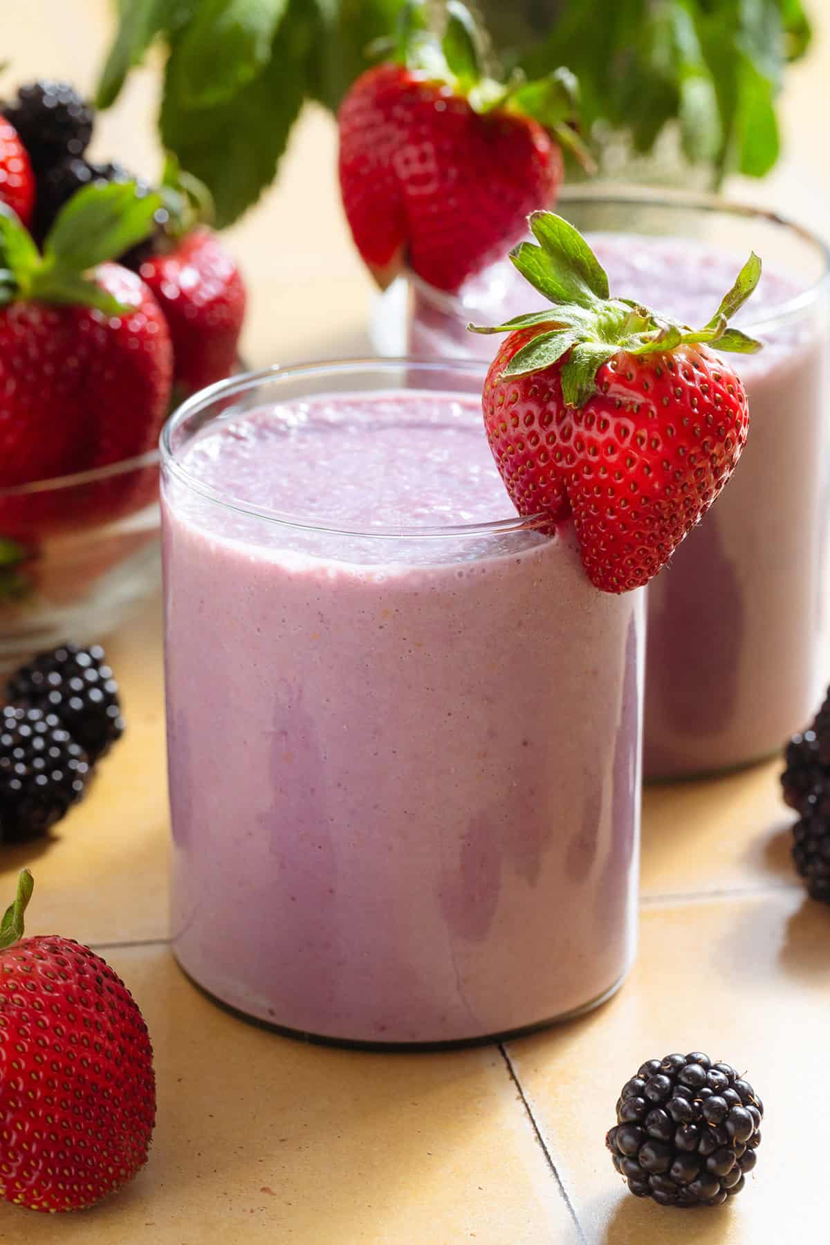 A light purple smoothie in a short glass garnished with a strawberry and blackberries around the glass.