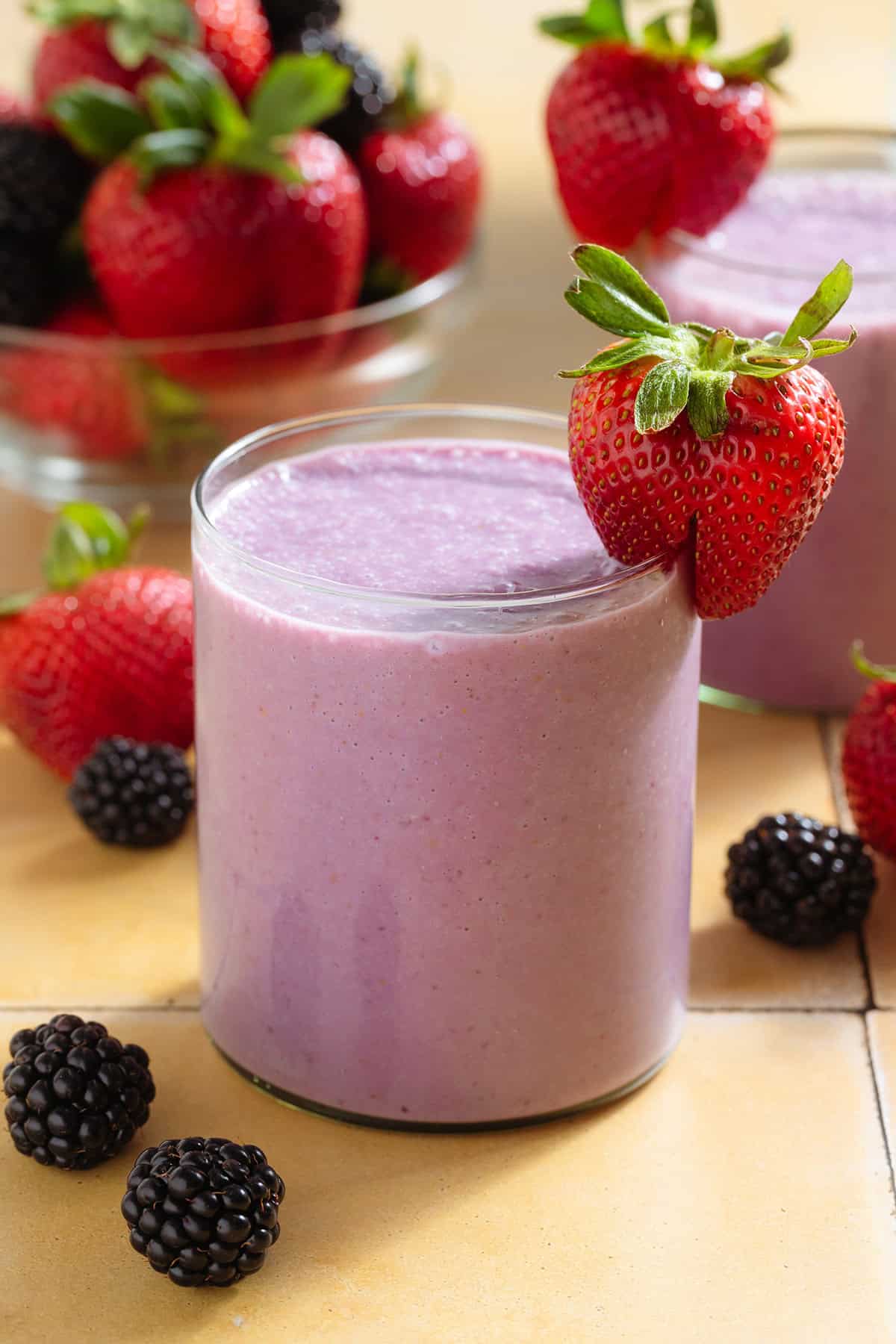A light purple smoothie in a short glass garnished with a strawberry and blackberries around the glass.
