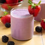 A light purple smoothie in a short glass garnished with a strawberry and blackberries around the glass.