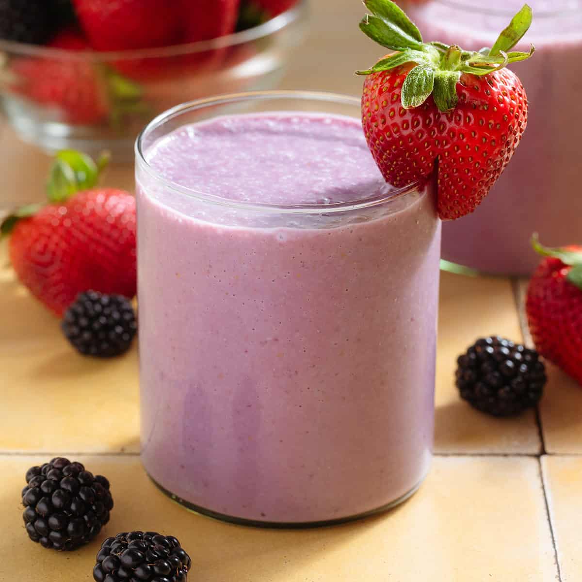 A light purple smoothie in a short glass garnished with a strawberry and blackberries around the glass.