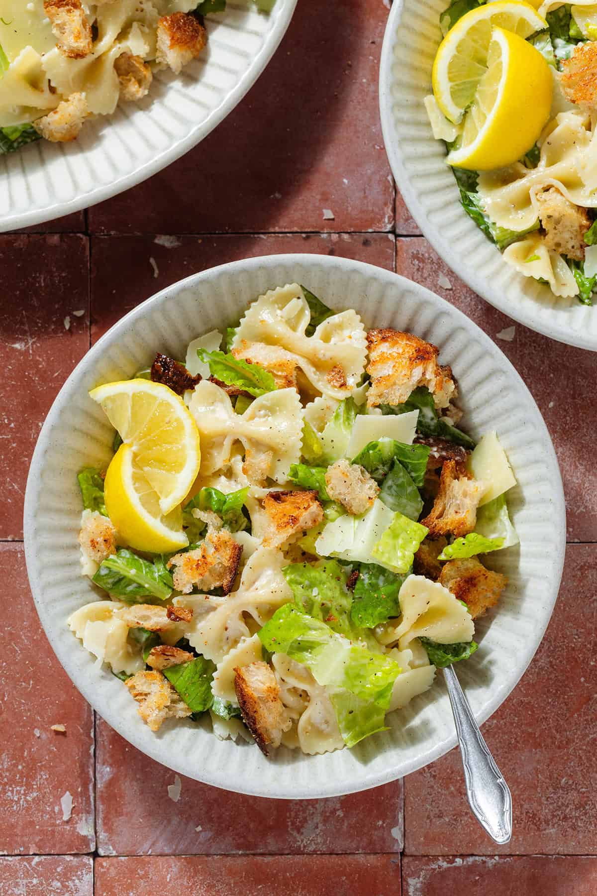 Caesar pasta salad with sourdough croutons in 3 low bowls, each garnished with two lemon wedges.