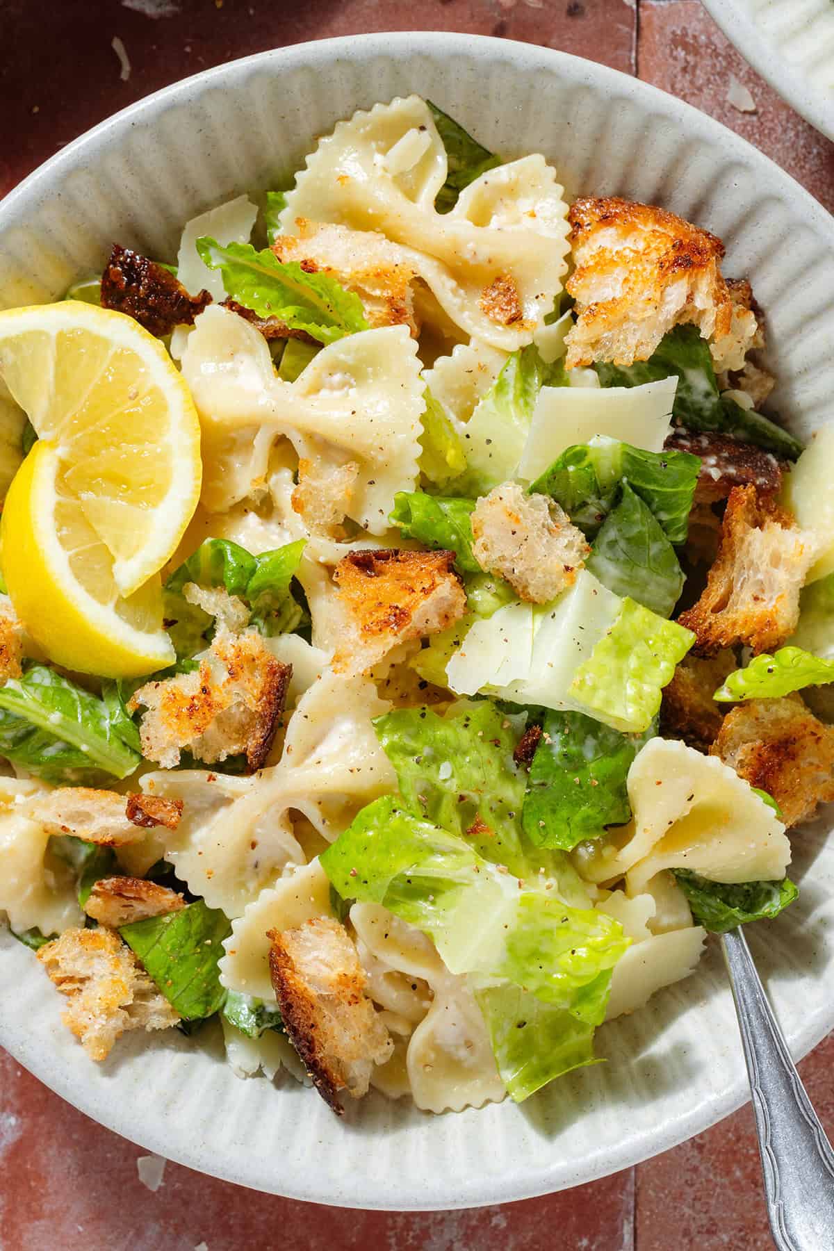 Caesar pasta salad with sourdough croutons in a low bowl garnished with two lemon wedges with a fork.