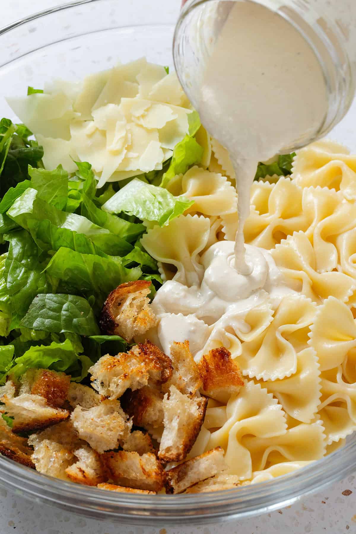 Caesar dressing being poured into a large bowl with chopped romaine lettuce, bow-tie pasta, croutons, and shaved parmesan.