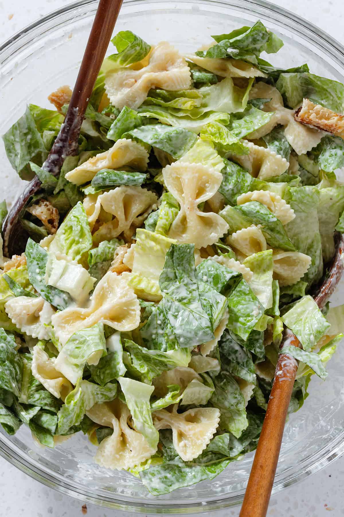 Chopped romaine lettuce, bow-tie pasta, and croutons being tossed with caesar dressing in a large glass bowl.