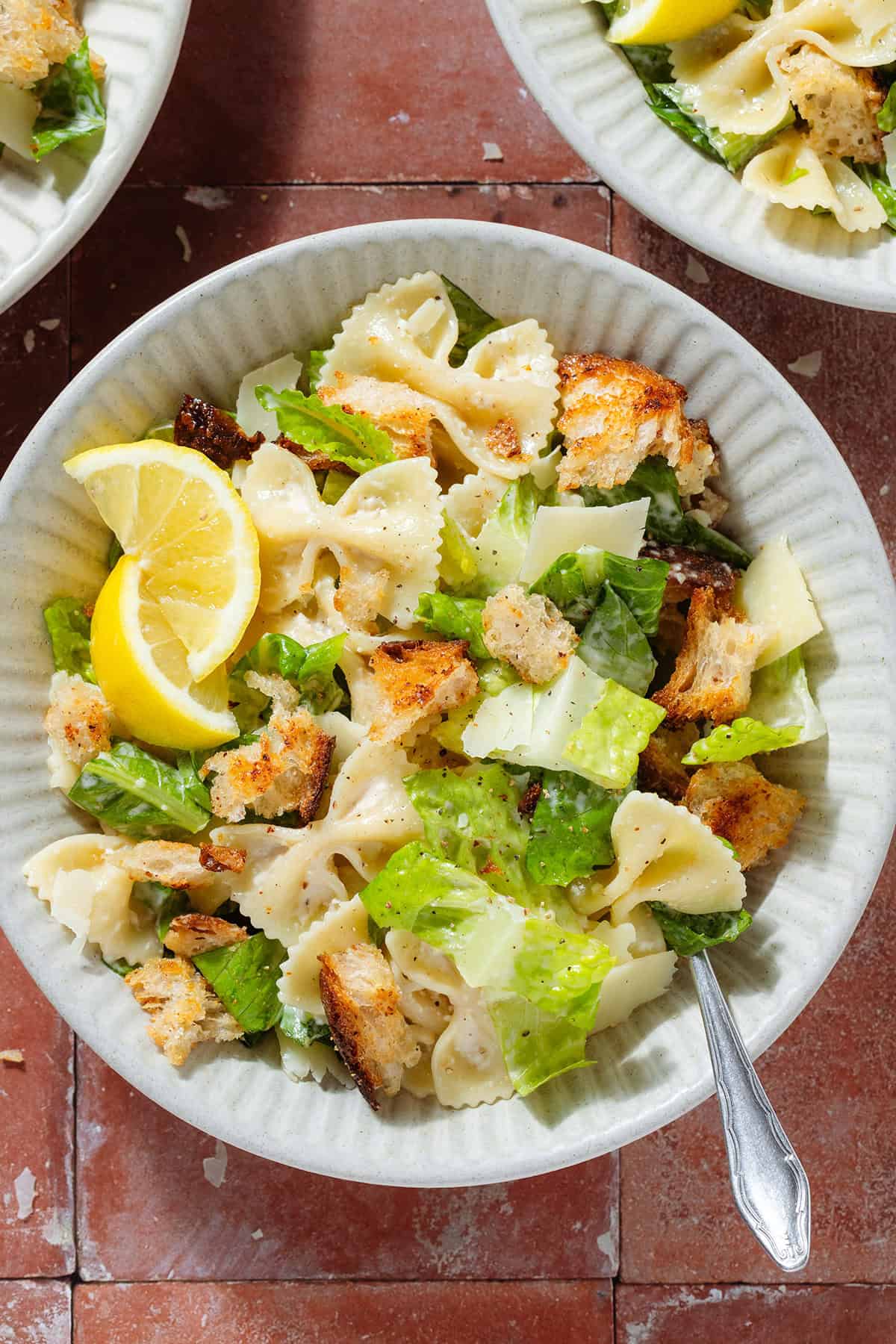 Caesar pasta salad with sourdough croutons in a low bowl garnished with two lemon wedges.
