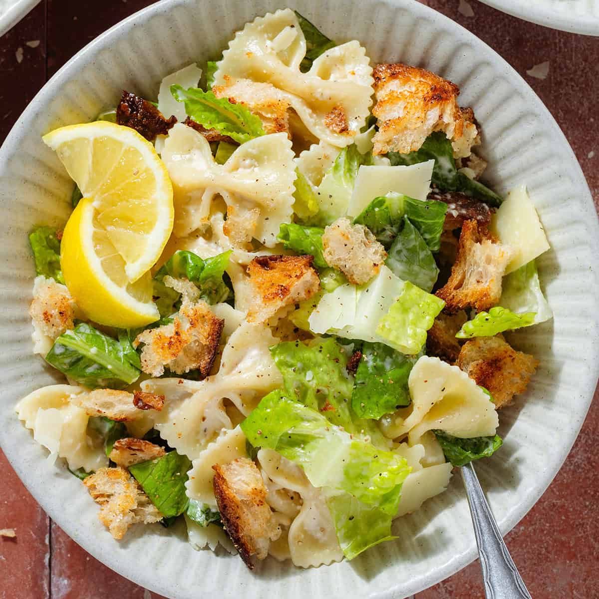 Caesar pasta salad with sourdough croutons in a low bowl garnished with two lemon wedges with a fork.