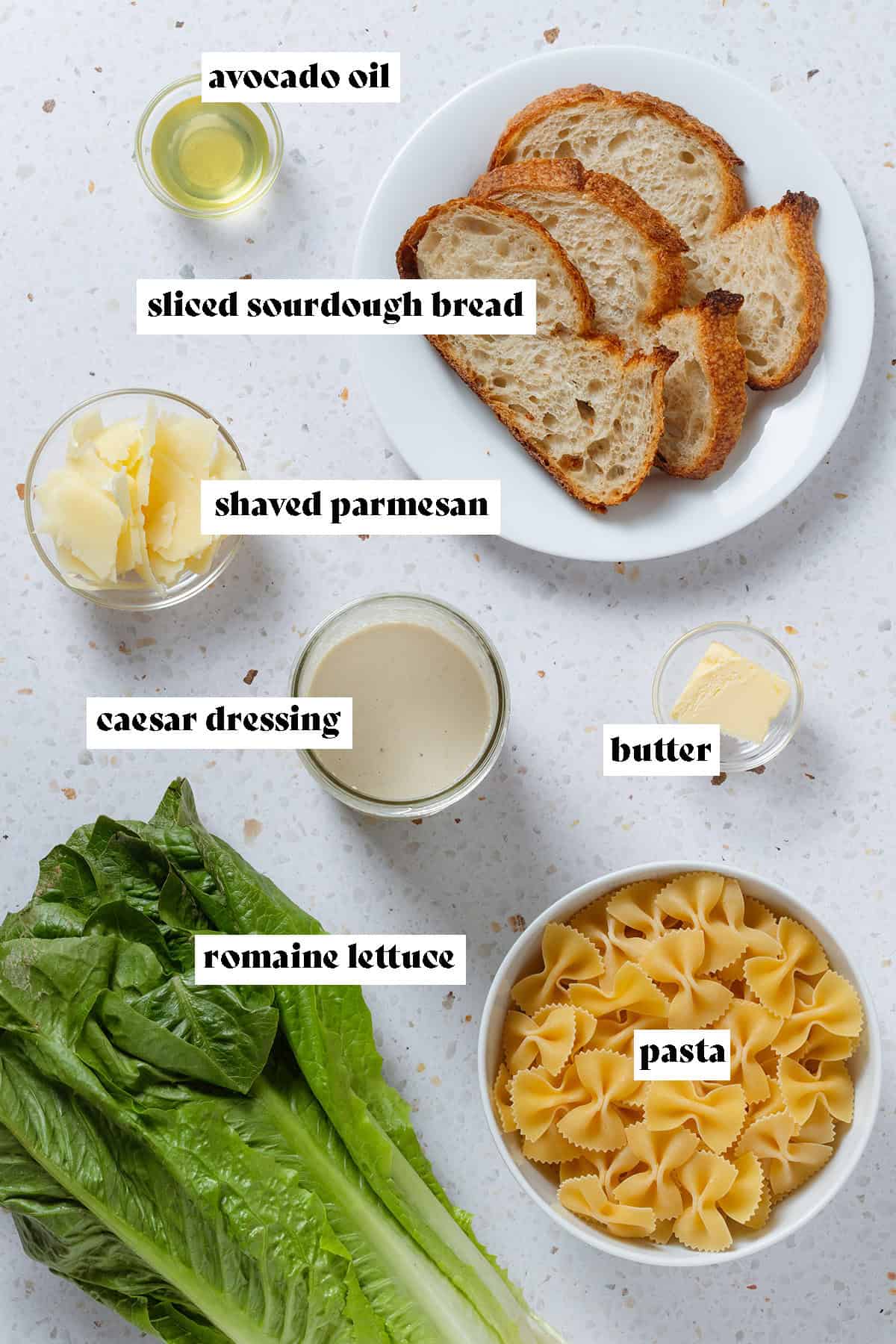 Sliced bread, dry pasta, romaine lettuce, caesar dressing, parmesan, oil, and butter in small bowls.