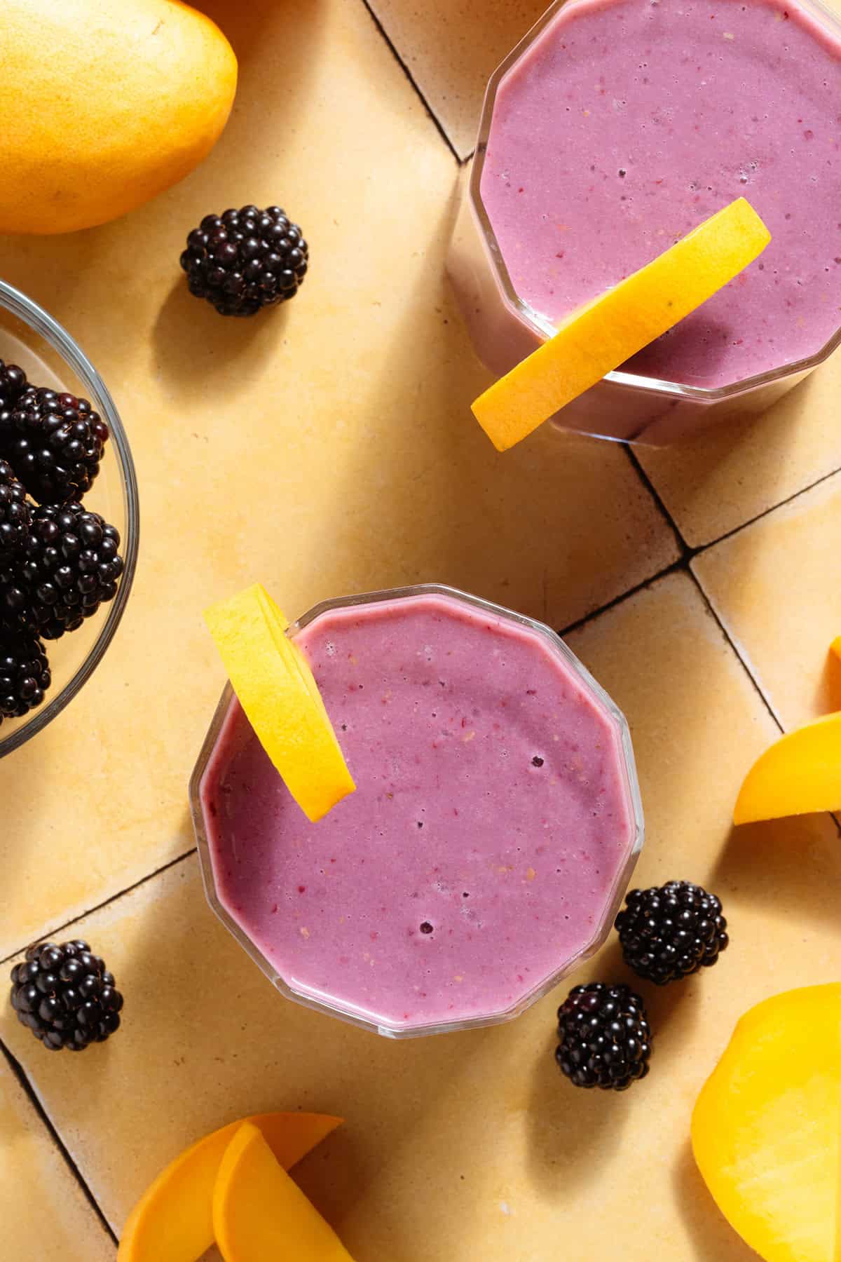 Vibrant purple smoothie in short glasses garnished with blackberries and slices of mango.
