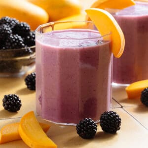Vibrant purple smoothie in short glasses garnished with blackberries and slices of mango.