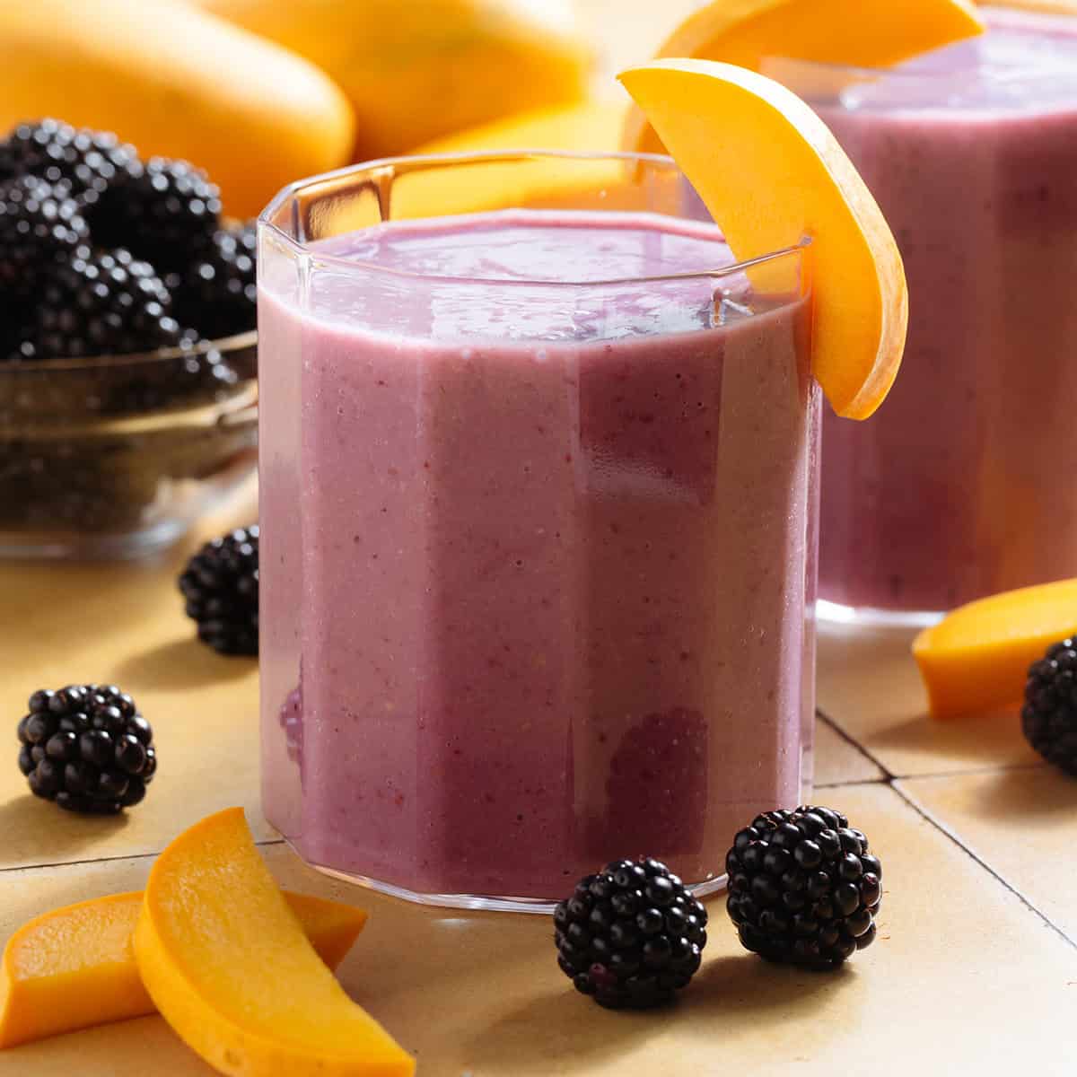 Vibrant purple smoothie in short glasses garnished with blackberries and slices of mango.