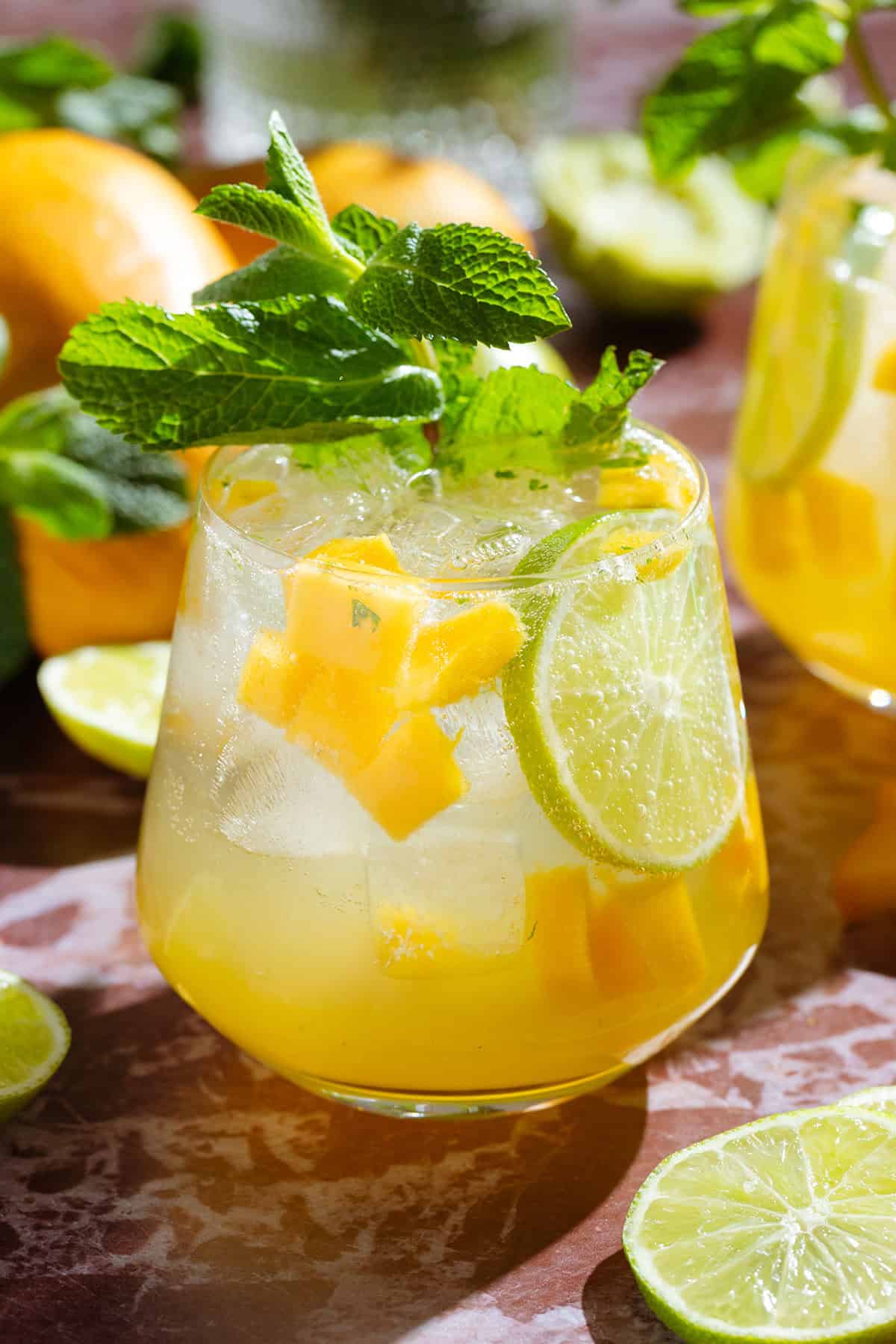 Bright yellow mango drink in a short glass garnished with fresh mint, a lime slice, and diced mango.