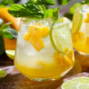 Bright yellow mango drink in a short glass garnished with fresh mint, a lime slice, and diced mango.