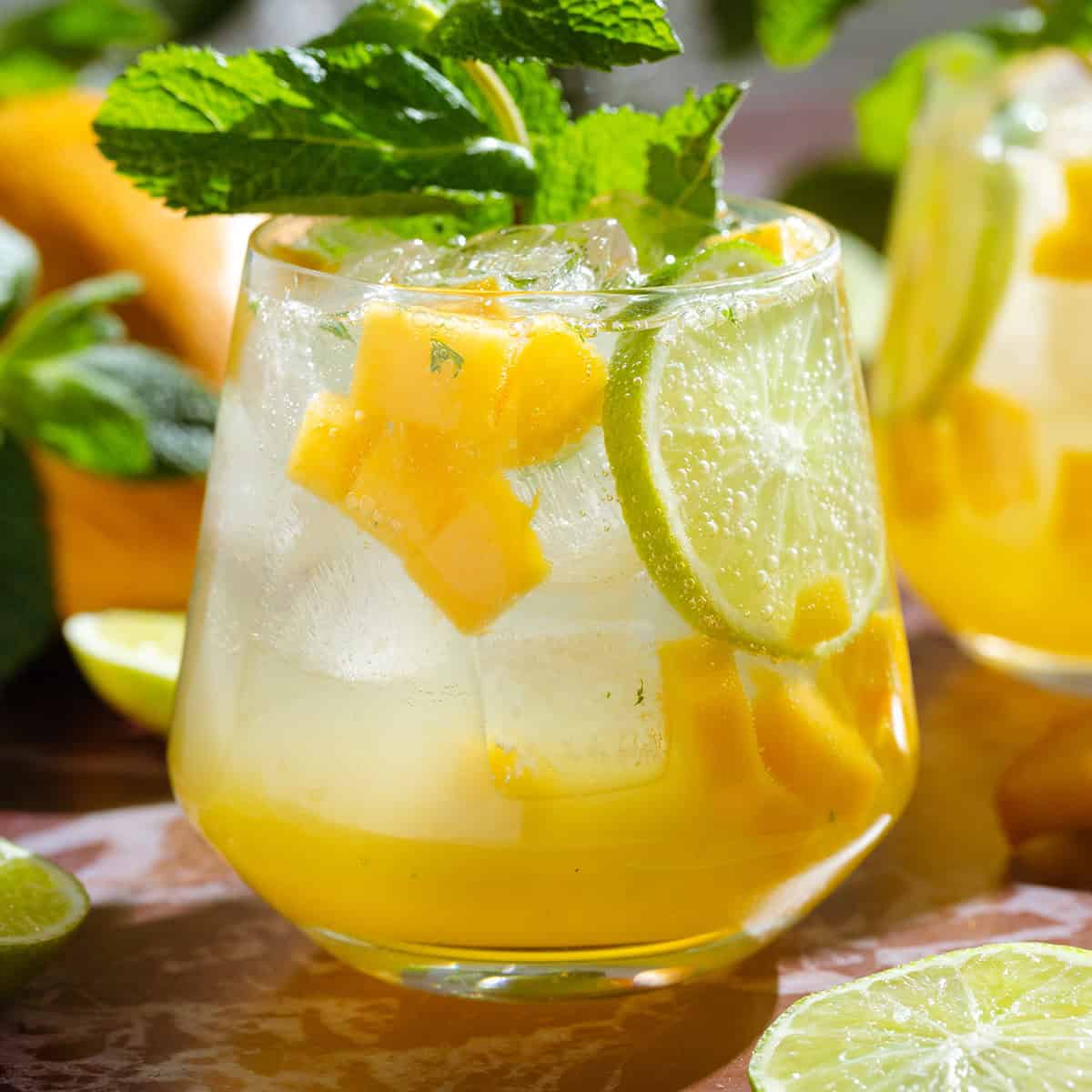 Bright yellow mango drink in a short glass garnished with fresh mint, a lime slice, and diced mango.