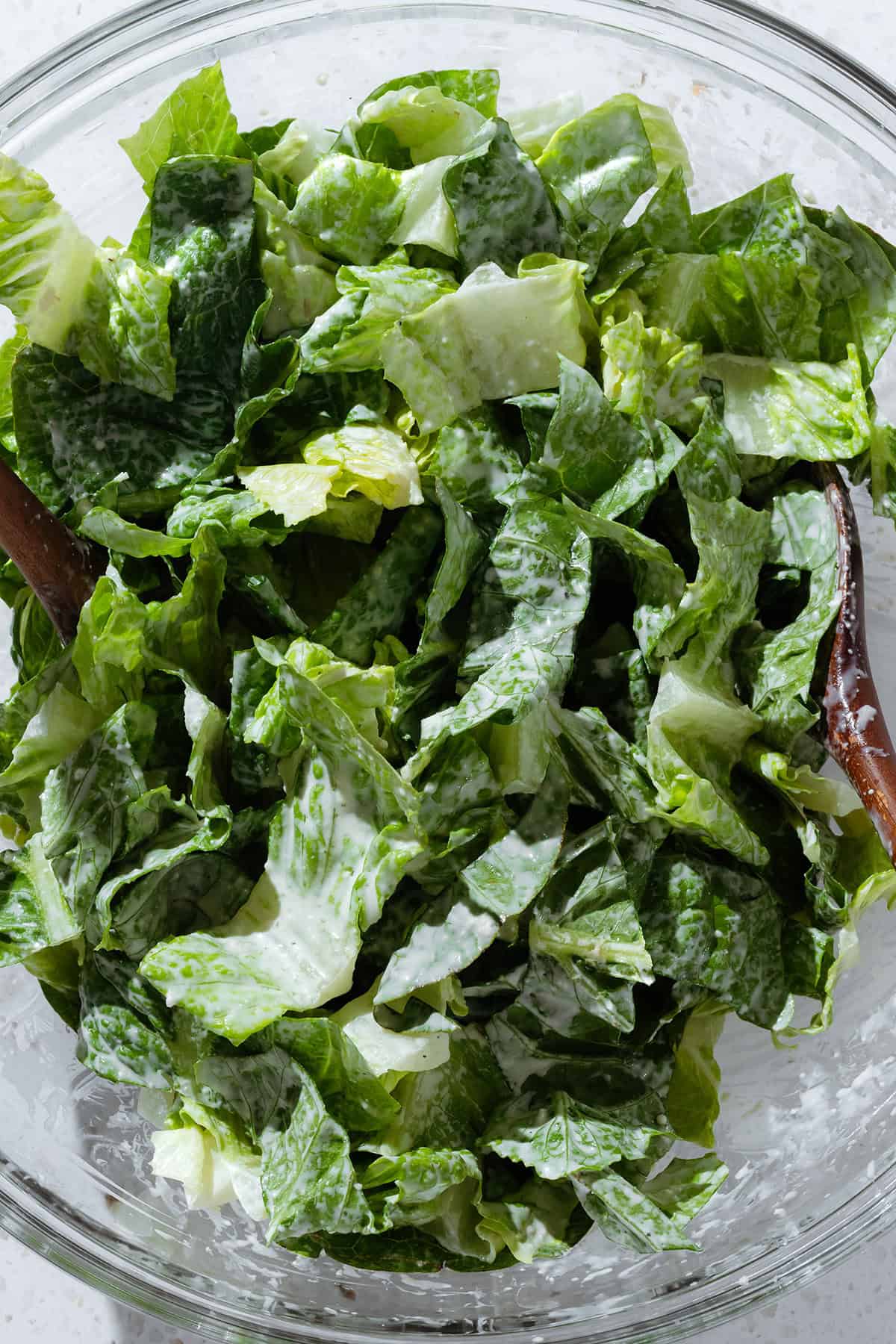 Caesar dressing tossed with chopped romaine lettuce in a large glass bowl with wooden serving spoons.