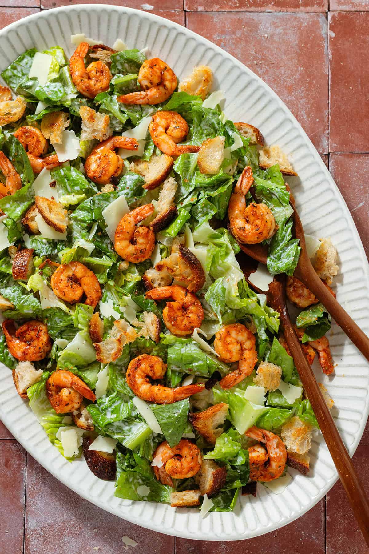 Caesar salad with spicy shrimp on a large oval serving platter with wooden serving spoons.