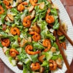 Caesar salad with spicy shrimp on a large oval serving platter with wooden serving spoons.