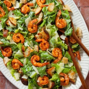 Caesar salad with spicy shrimp on a large oval serving platter with wooden serving spoons.