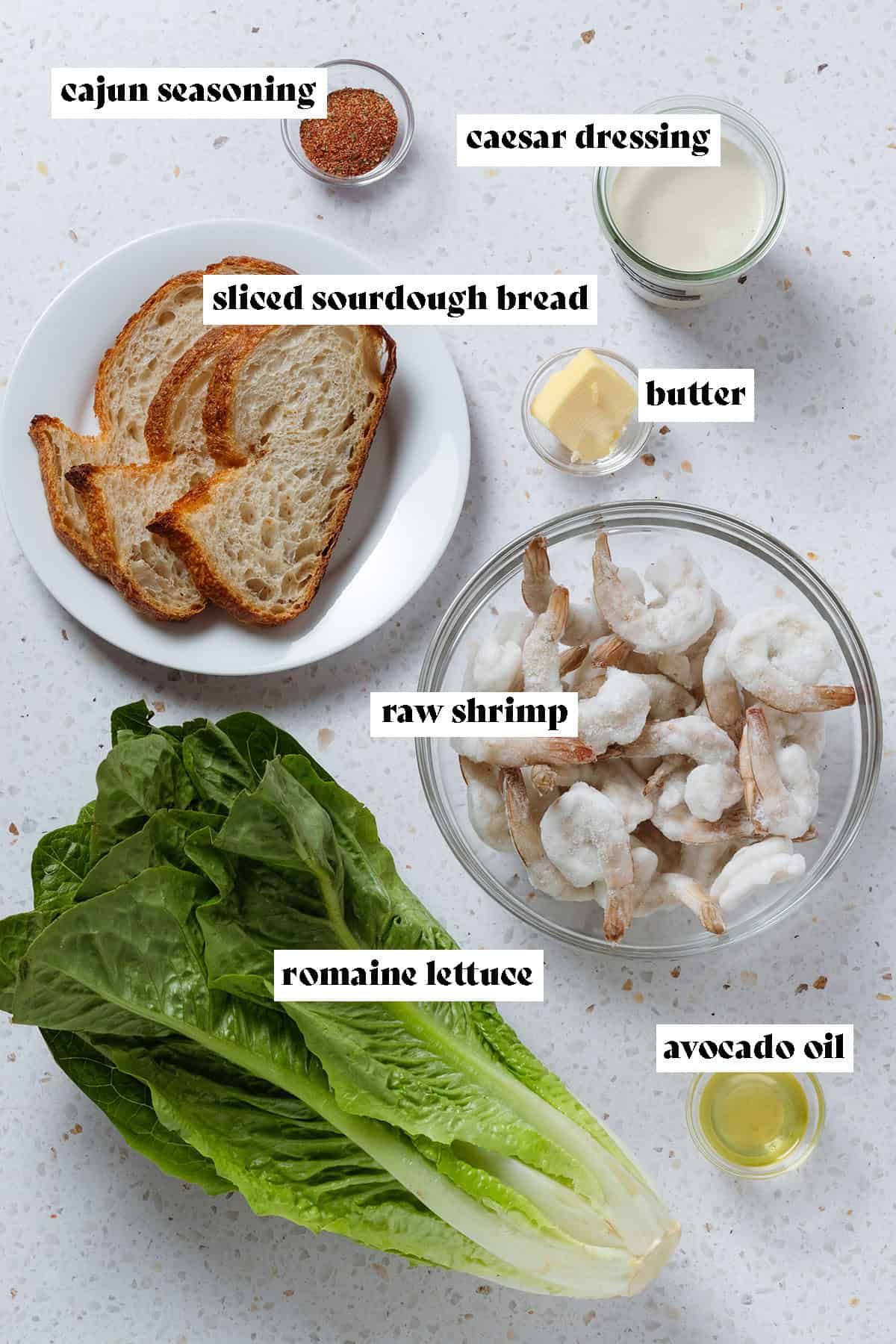 Romaine lettuce, raw shrimp, slices of bread, dressing, and other ingredients laid out on a stone background.