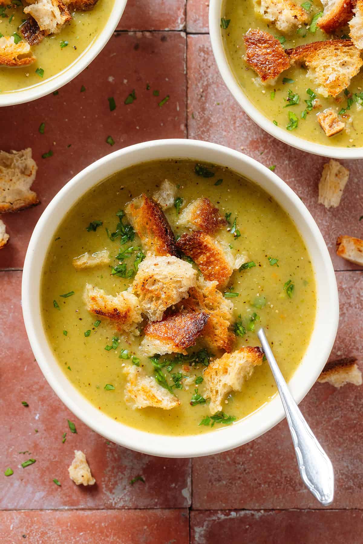 Creamy zucchini soup topped with sourdough croutons and chopped fresh parsley in a white bowl with a spoon in the soup on the right.