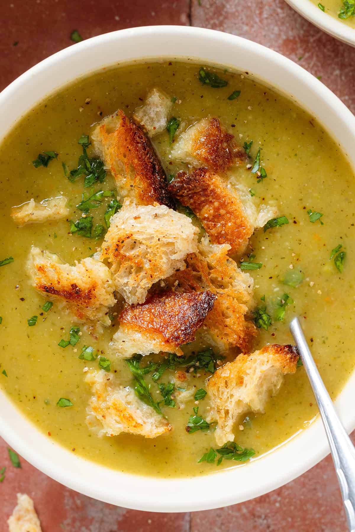 Creamy zucchini soup topped with sourdough croutons and chopped fresh parsley in a white bowl with a spoon in the soup on the right.