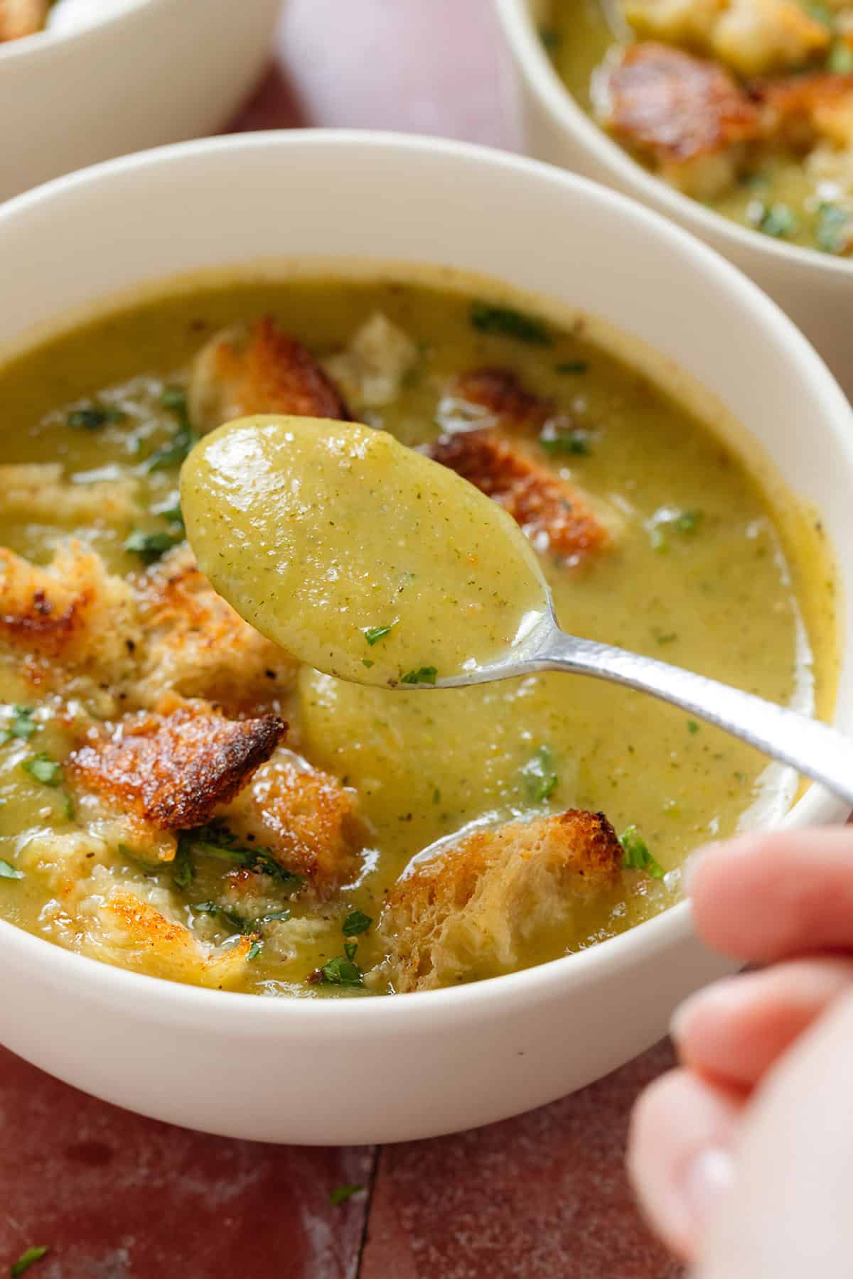 Creamy zucchini soup topped with sourdough croutons and chopped fresh parsley in a white bowl with a spoon picking up the soup.