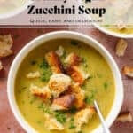 Creamy zucchini soup topped with sourdough croutons and chopped fresh parsley in a white bowl with a spoon in the soup on the right.