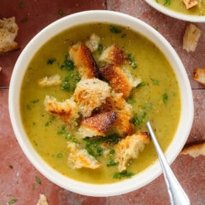 Creamy zucchini soup topped with sourdough croutons and chopped fresh parsley in a white bowl with a spoon in the soup on the right.