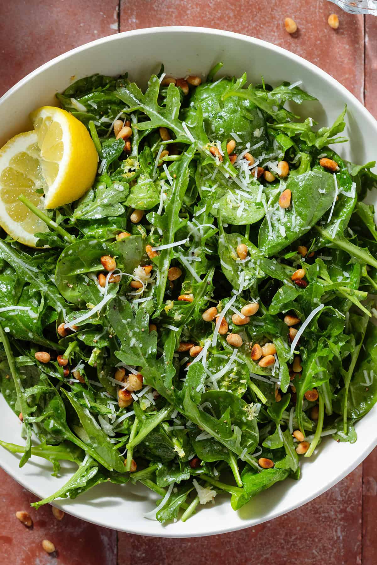A large bowl with arugula spinach salad sprinkled with grated parmesan and toasted pine nuts with two lemon wedges.