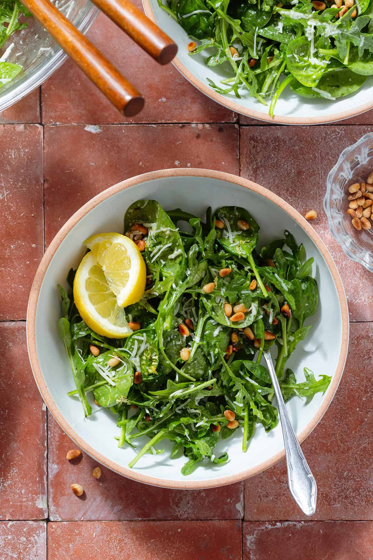 Spinach arugula salad sprinkled with parmesan and toasted pine nuts with two lemon wedges on the side in two bowls with forks.