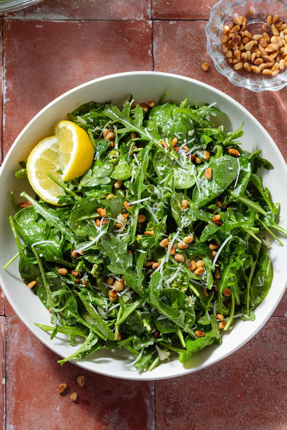 A large bowl with arugula spinach salad sprinkled with grated parmesan and toasted pine nuts with two lemon wedges.