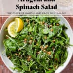 A large bowl with arugula spinach salad sprinkled with grated parmesan and toasted pine nuts with two lemon wedges.