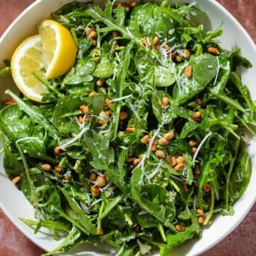 A large bowl with arugula spinach salad sprinkled with grated parmesan and toasted pine nuts with two lemon wedges on the left.
