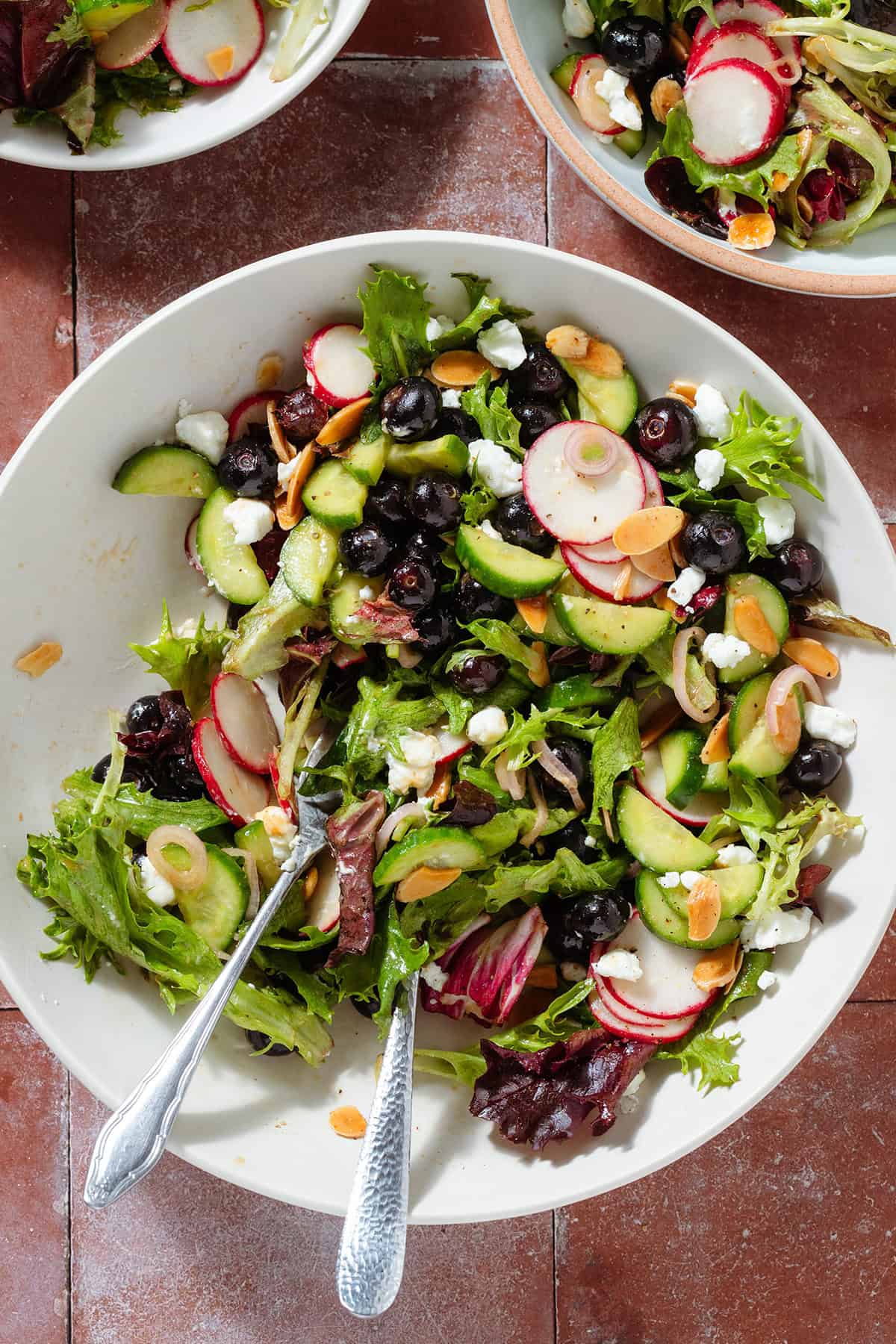 A half empty bowl on blueberry salad with mixed greens, radishes, toasted almonds, shallots, and crumbled goat cheese with serving forks on the left.