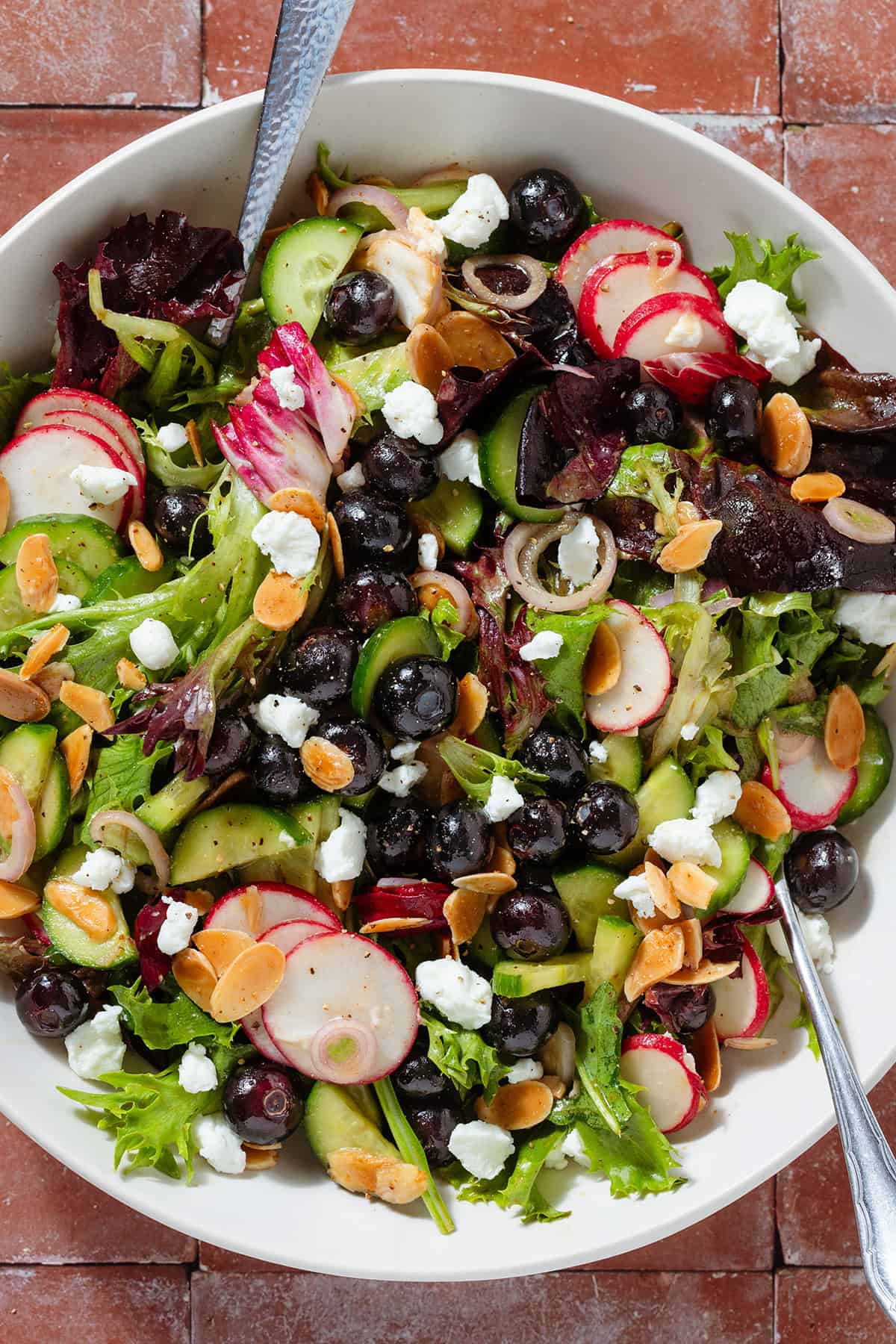 A bowl on blueberry salad with mixed greens, radishes, toasted almonds, goat cheese, and shallots with serving spoons.