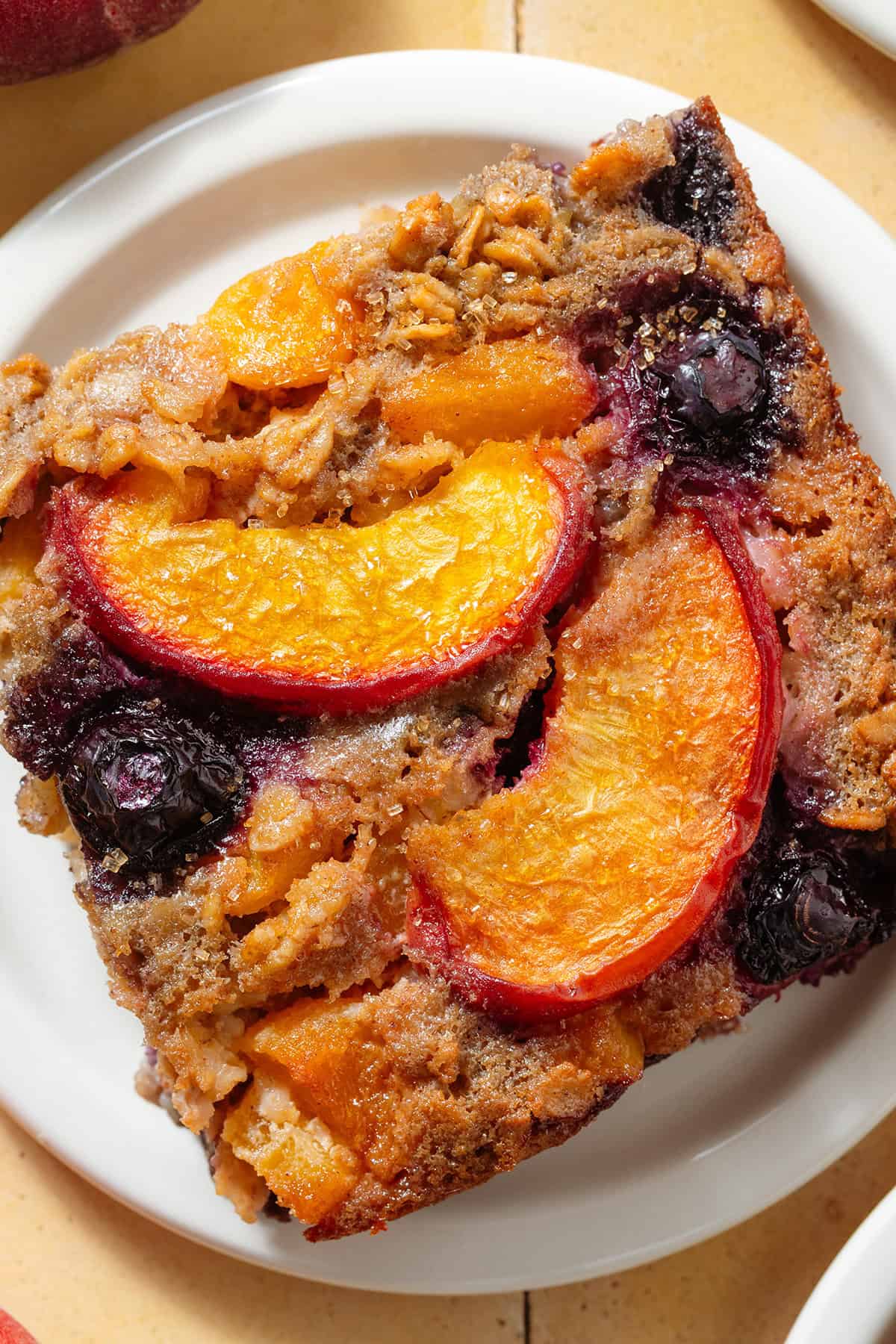 A baked oatmeal square with blueberries and peach slices and a caramelized top on small white plate.
