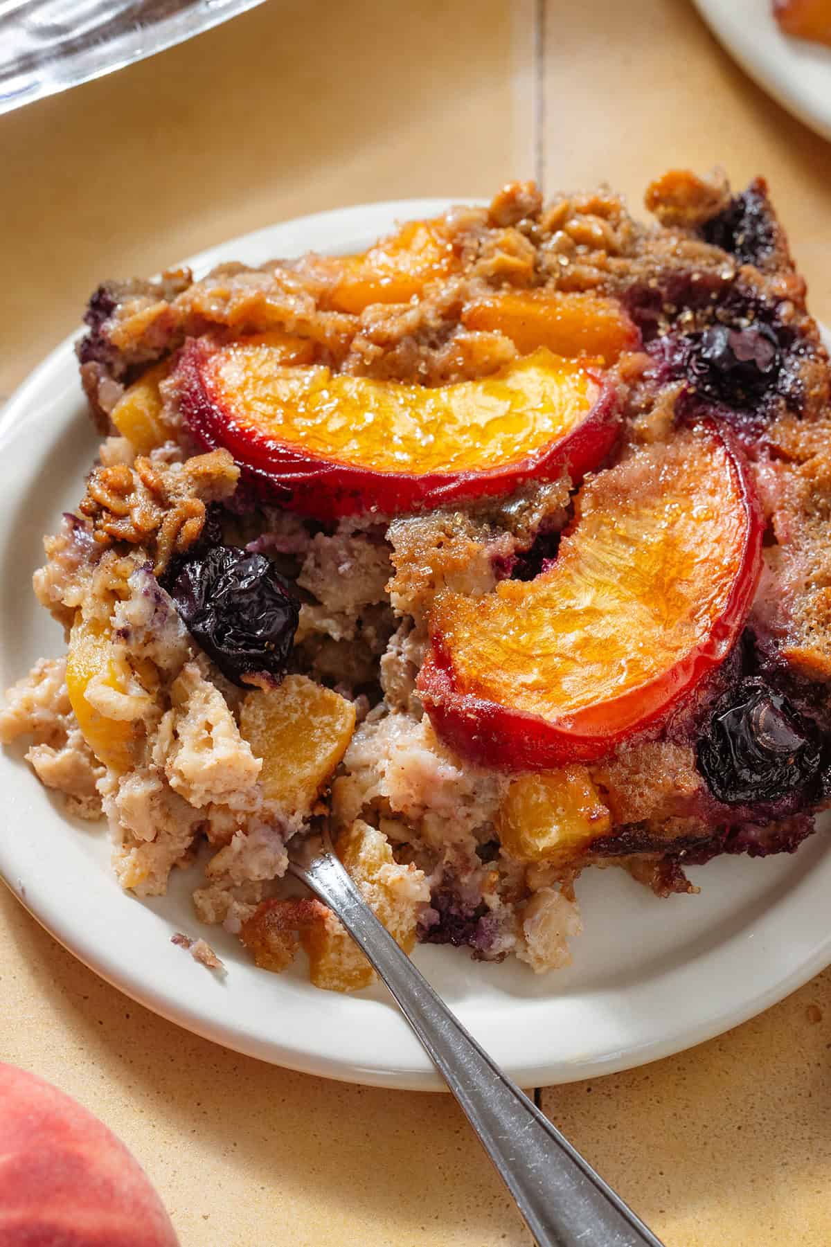 A baked oatmeal square with caramelized blueberries and peach slices in it on small white plate.