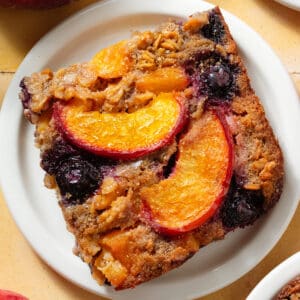 A baked oatmeal square with caramelized blueberries and peach slices in it on small white plate.