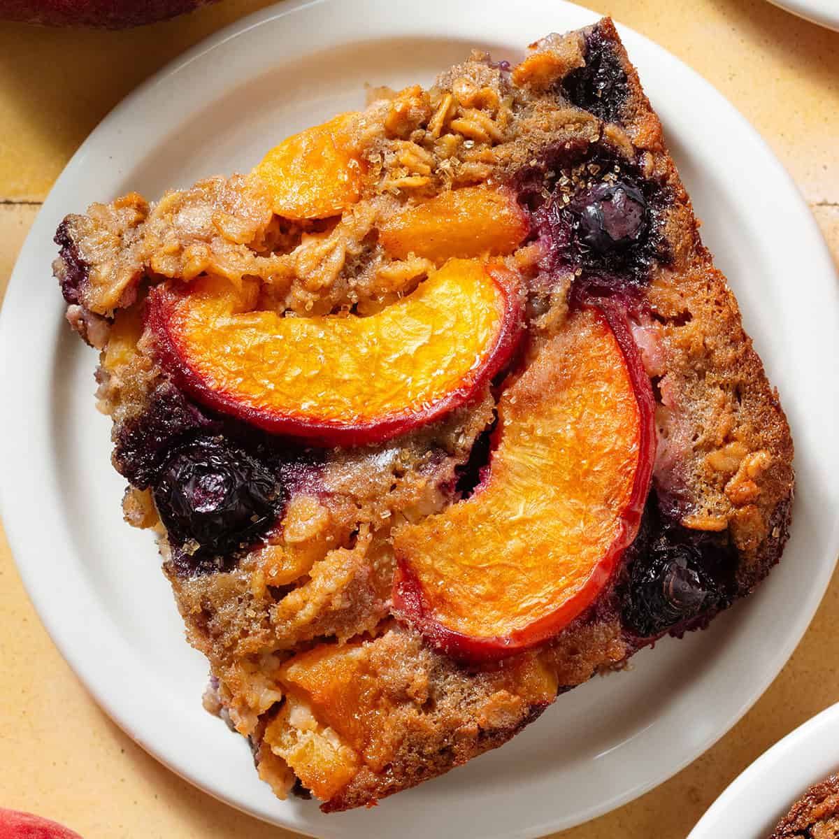 A baked oatmeal square with caramelized blueberries and peach slices in it on small white plate.