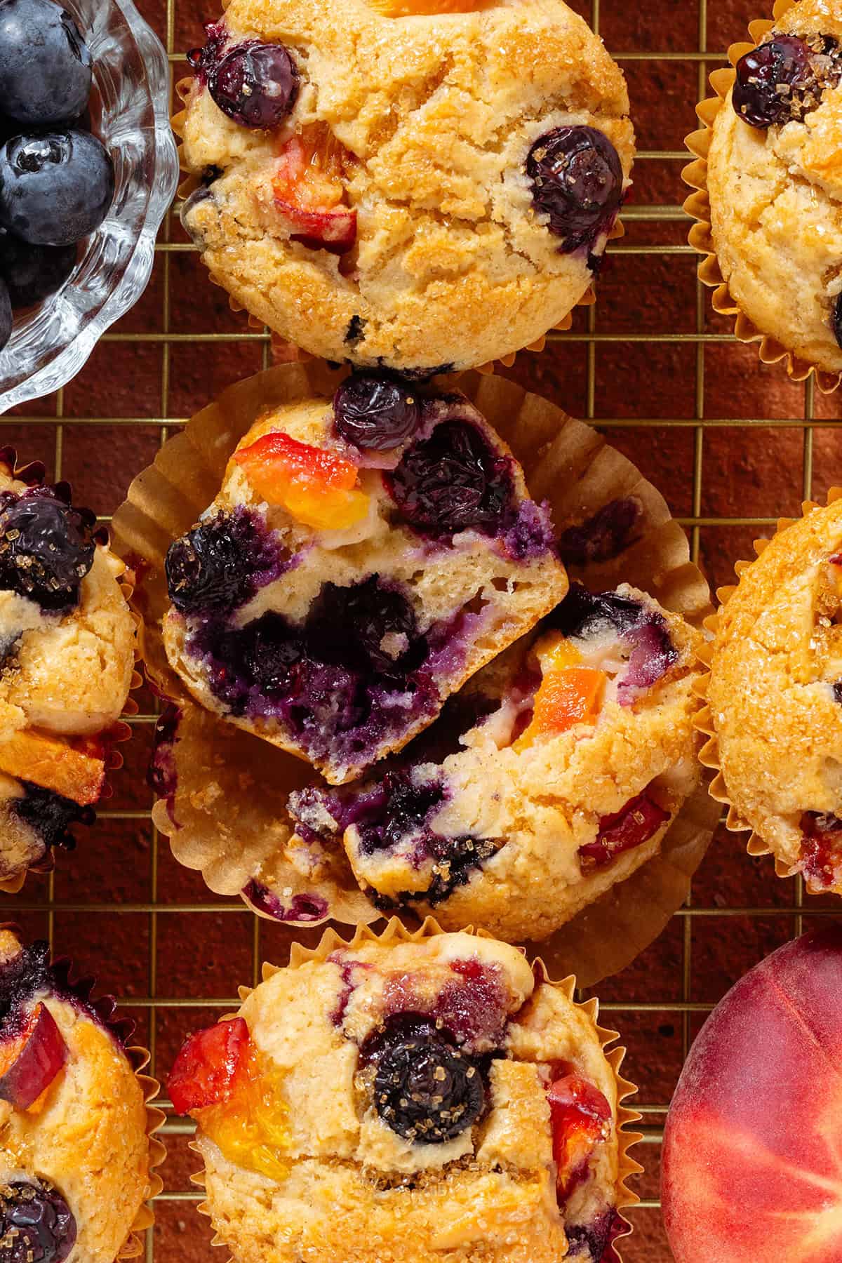 Blueberry peach muffins on a golden cooling wire rack with the muffin in the middle sliced in half to show the melted blueberries inside.