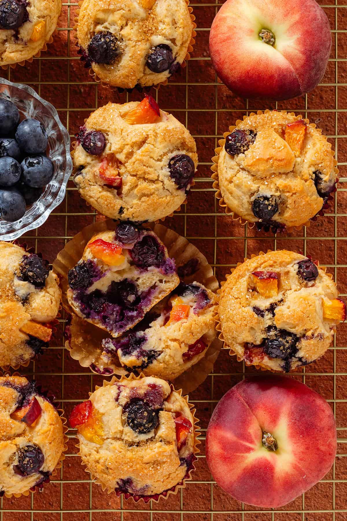 Blueberry peach muffins on a golden cooling wire rack with the muffin in the middle sliced in half to show the melted blueberries inside with fresh blueberries and peaches in between them.