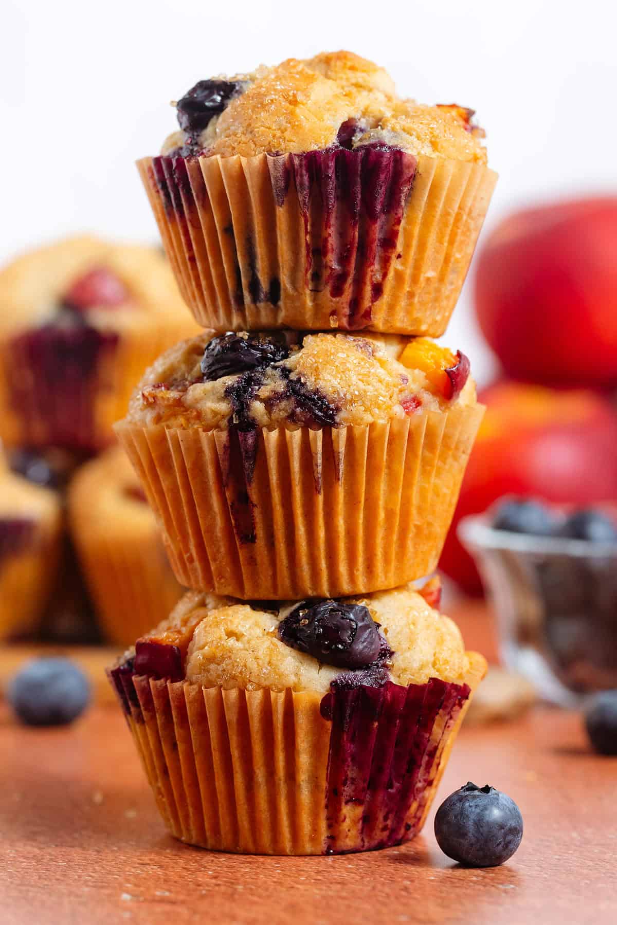 Three blueberry peach muffins stacked with more muffins, peaches, and blueberries behind them.