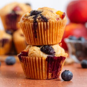 Two blueberry peach muffins stacked with more muffins, peaches, and blueberries behind them.