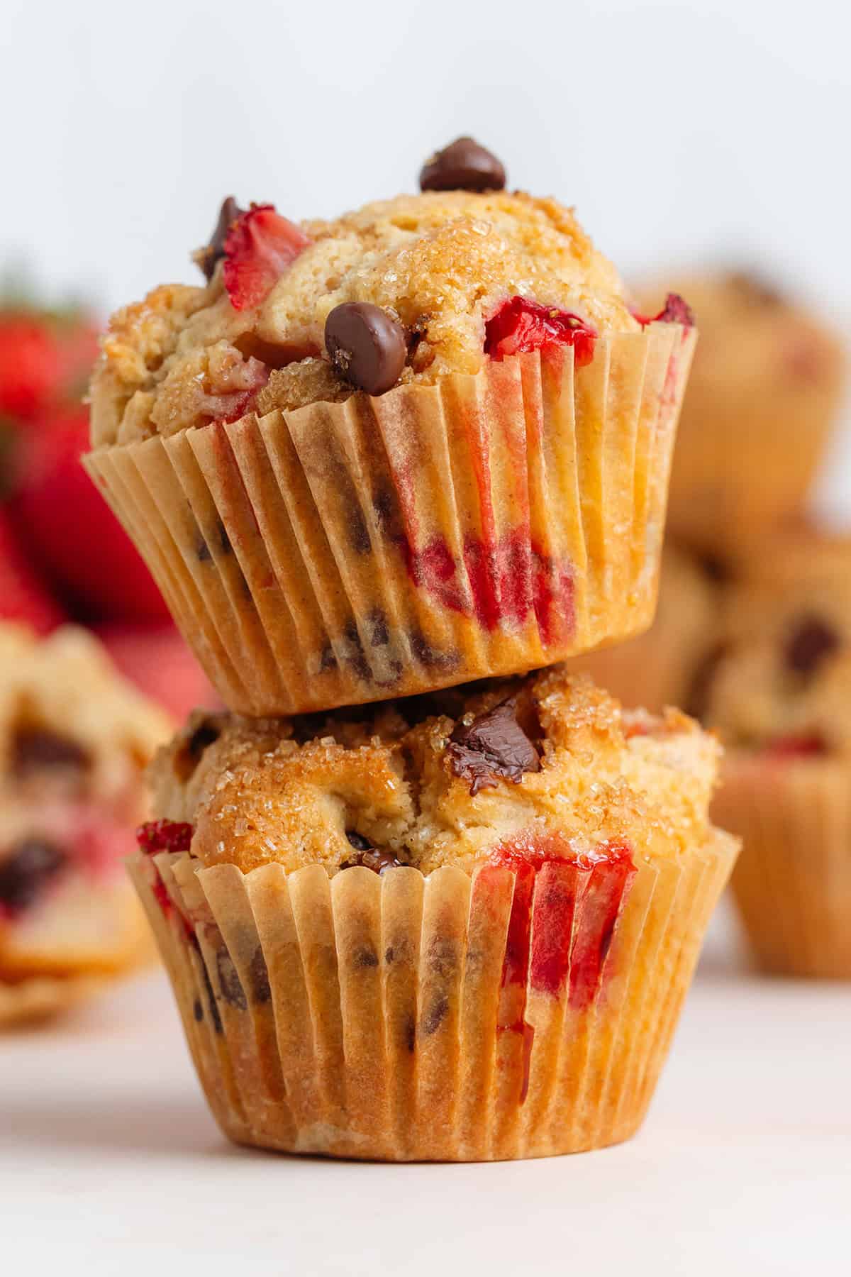 Two muffins with strawberries and chocolate chips stacked on a light pink background with more muffins in the back.